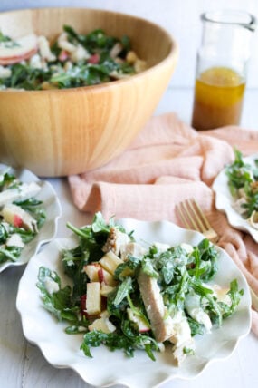 A plate of turkey arugula salad with chicken, apples, and cheese up front; big bowl and dressing jar in back, pink napkin by the plate.