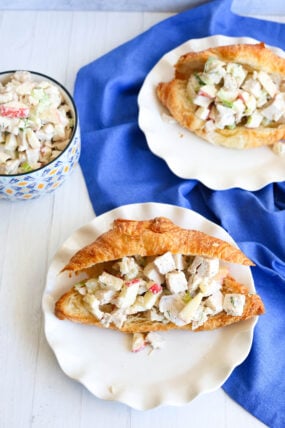 Two croissant sandwiches stuffed with chicken or turkey salad (with apples and celery) sit on plates. Extra salad nearby.
