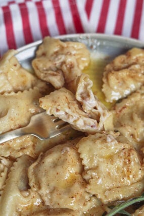 Creamy brie ravioli with buttery sauce and spices sits on a plate. A fork shows the cheesy middle. Red striped cloth in back.