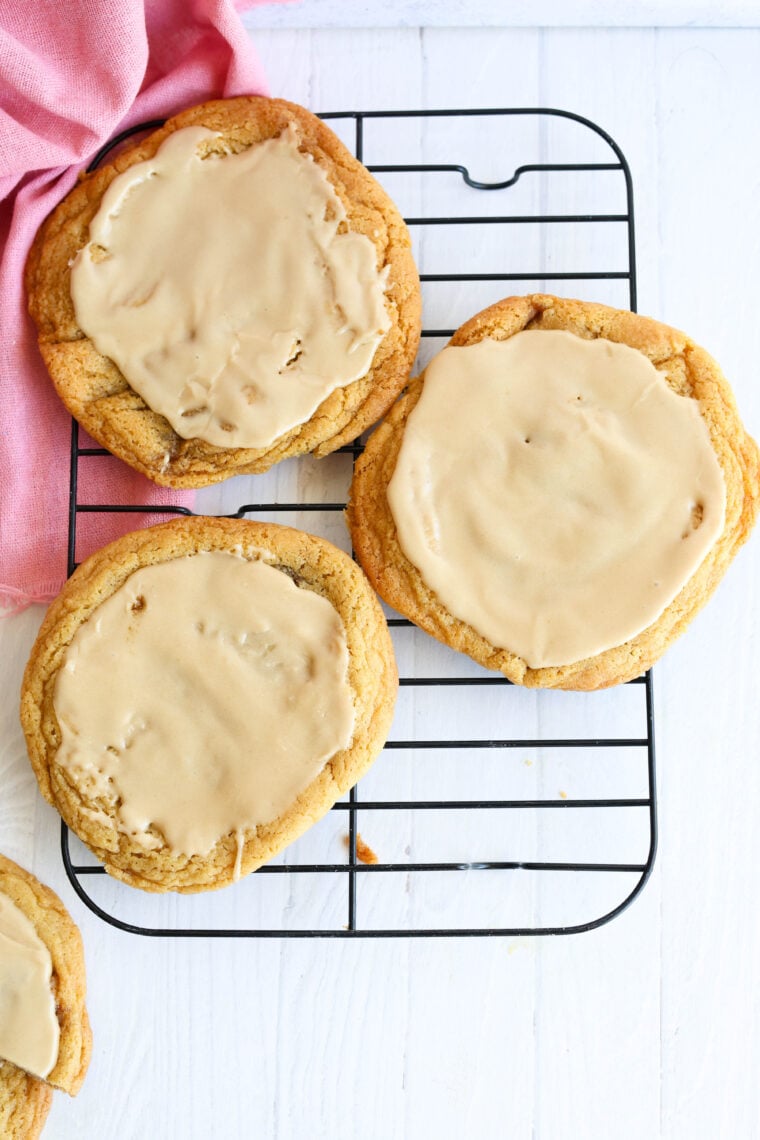 Three big cookies with brown sugar inside and shiny maple icing chill on a black rack; a pink cloth peeks in from the corner.