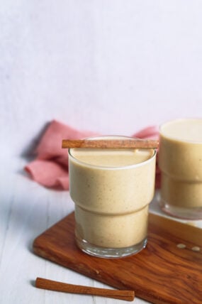 A glass of creamy eggnog smoothie with a cinnamon stick sits on a wooden board, plus another glass and pink cloth in back.