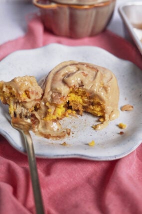 A half-eaten pumpkin cinnamon roll with frosting sits on a white plate, fork nearby, all on a soft pink cloth.