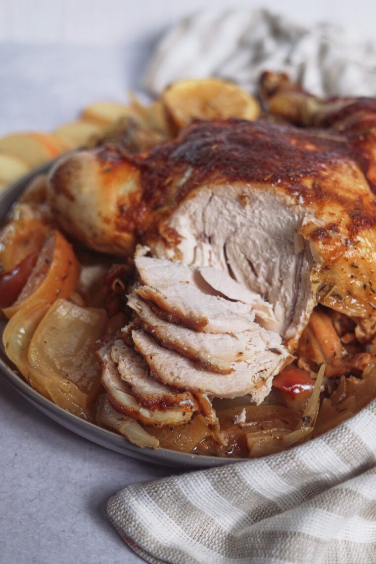 A golden roasted chicken sits on a platter with sliced meat, onions, apples, and potatoes, plus a striped towel off to the side.
