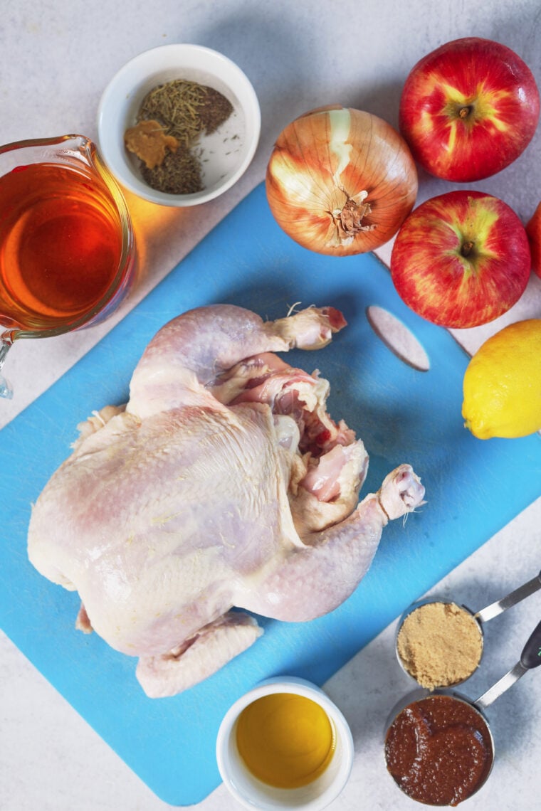 A raw chicken sits on a blue board with apples, onion, lemon, brown sugar, spices, oil, sauce, and a cup of liquid nearby.