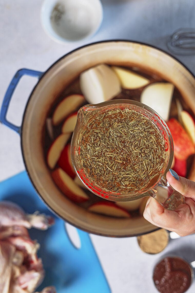 Someone’s about to cook, holding dried herbs over a pot with apples and onions. A roast chicken, spices, and bowl chill nearby.