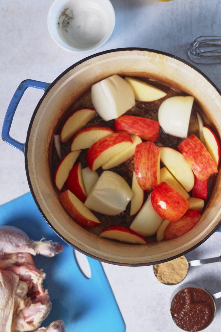 A big pot with chopped apples and onions is on the counter, next to a roast chicken, some spices, sauce, and a small bowl.