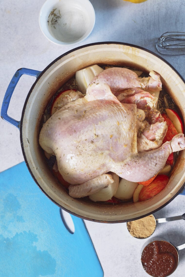 A roasted chicken in a blue Dutch oven with apples and onions, plus a white bowl, blue cutting board, brown sugar, and BBQ sauce.
