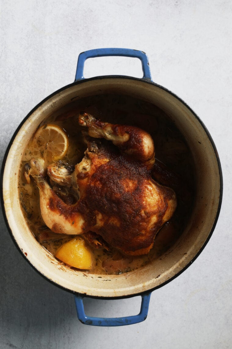 Overhead shot of a golden roasted chicken in a blue Dutch oven, with lemon slices and herbs all around, on a light gray table.