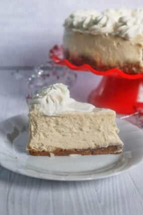 A piece of eggnog cheesecake with whipped cream and a graham crust sits on a plate, more cheesecake on a red stand behind.