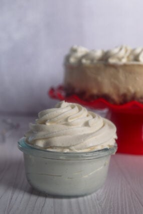 A little glass bowl of fluffy, pale eggnog whipped cream up front, with a swirly-frosted cake on a red stand behind it.