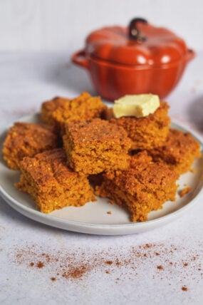 Pumpkin cornbread squares with melty butter on a plate, sitting by a pumpkin dish. Crumbs and cinnamon are scattered around.
