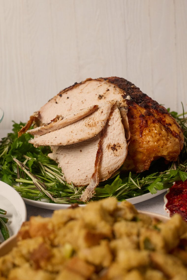 Sliced roast turkey breast on fresh herbs, with stuffing and cranberry sauce up front—perfectly thawed and ready for dinner!.