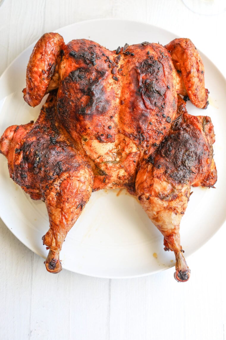 A crispy, golden spatchcock chicken with tasty seasonings sits on a white plate on a white wooden table, looking delicious.