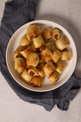A white bowl packed with pretzel-wrapped pigs in a blanket sits on a dark gray cloth, all on a light surface.