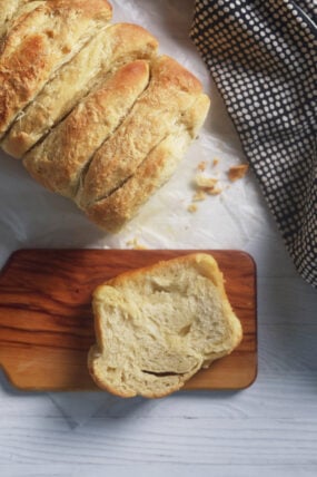 A crusty sourdough loaf sits on parchment next to a black-and-white cloth, with a slice on a small wooden board.