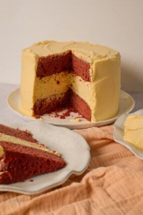 Red velvet cheesecake with yellow frosting sits on a white table&mdash;slice out, layers showing off, peach cloth nearby.