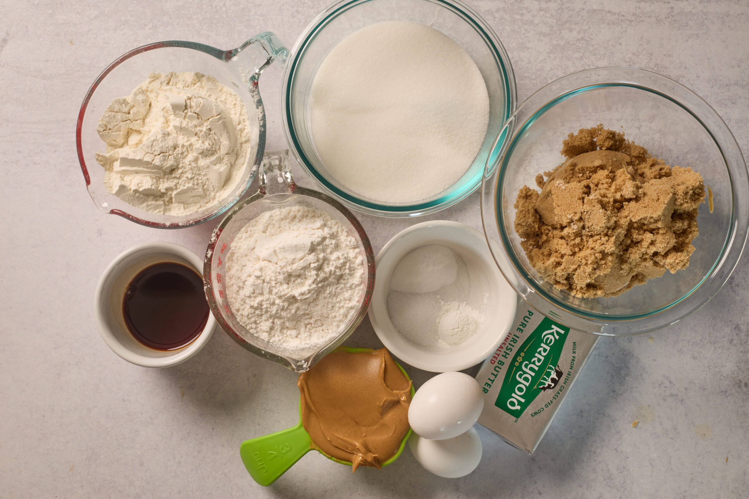 Top-down shot of flour, sugars, vanilla, peanut butter, eggs, butter, and baking powders in bowls on a light counter.