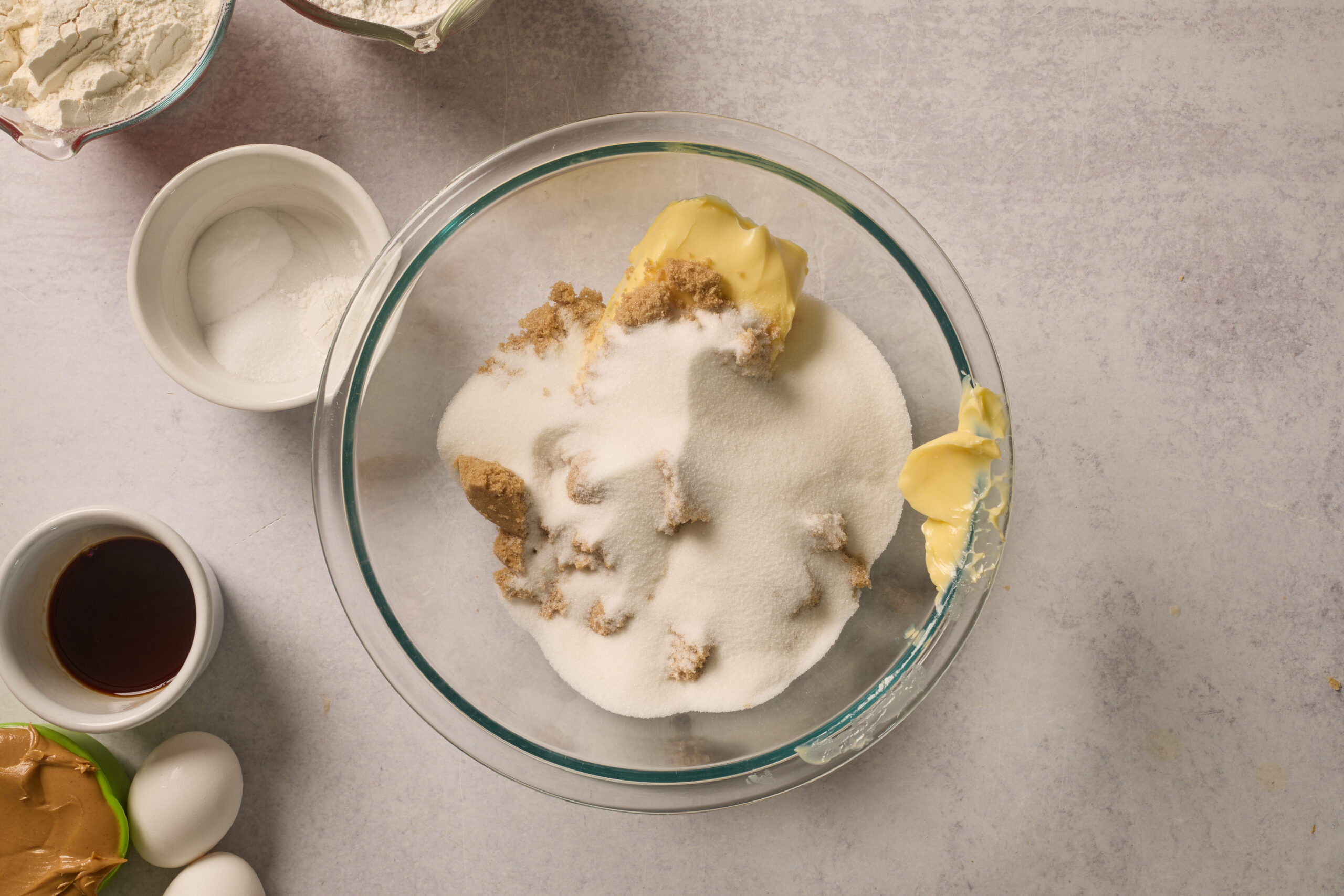 Big glass bowl with sugar and butter sits in the middle, surrounded by bowls of flour, salt, vanilla, peanut butter, and eggs.
