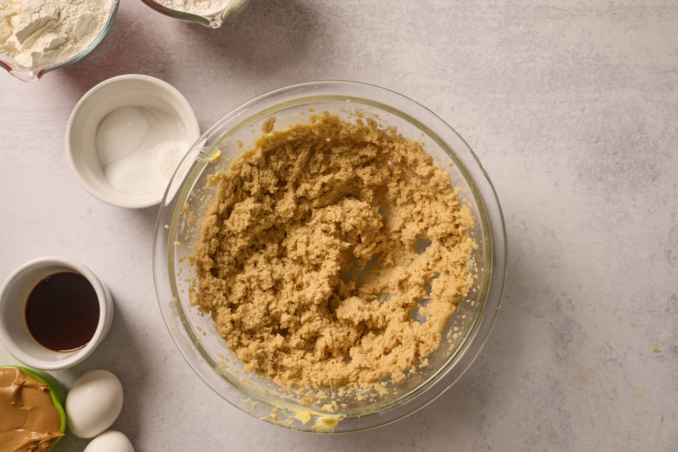 A glass bowl of half-mixed cookie dough on the counter, with flour, sugar, vanilla, eggs, and peanut butter bowls around it.