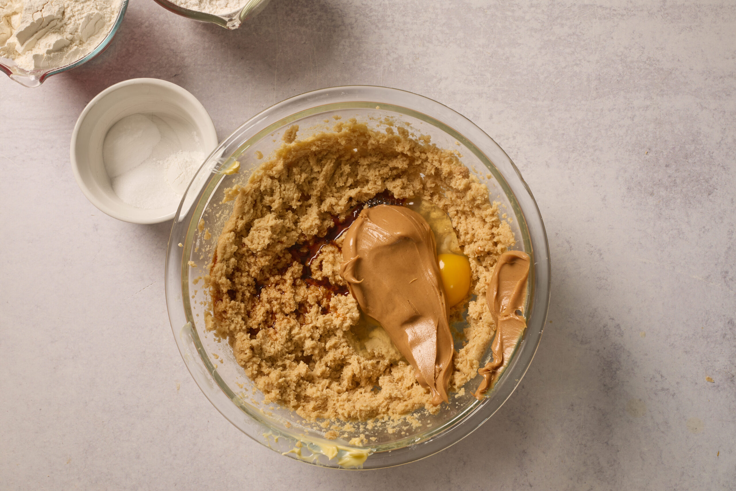 A glass bowl with brown sugar, an egg, vanilla, and peanut butter sits by bowls of flour and baking soda on a light counter.