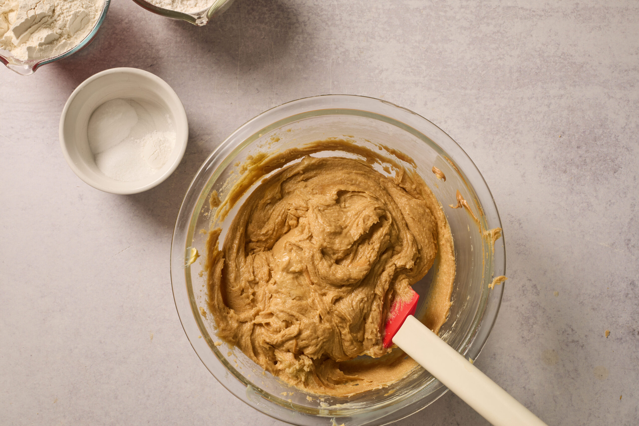A glass bowl full of cookie dough and a spatula sits next to some flour and a little bowl of white powder on a light counter.