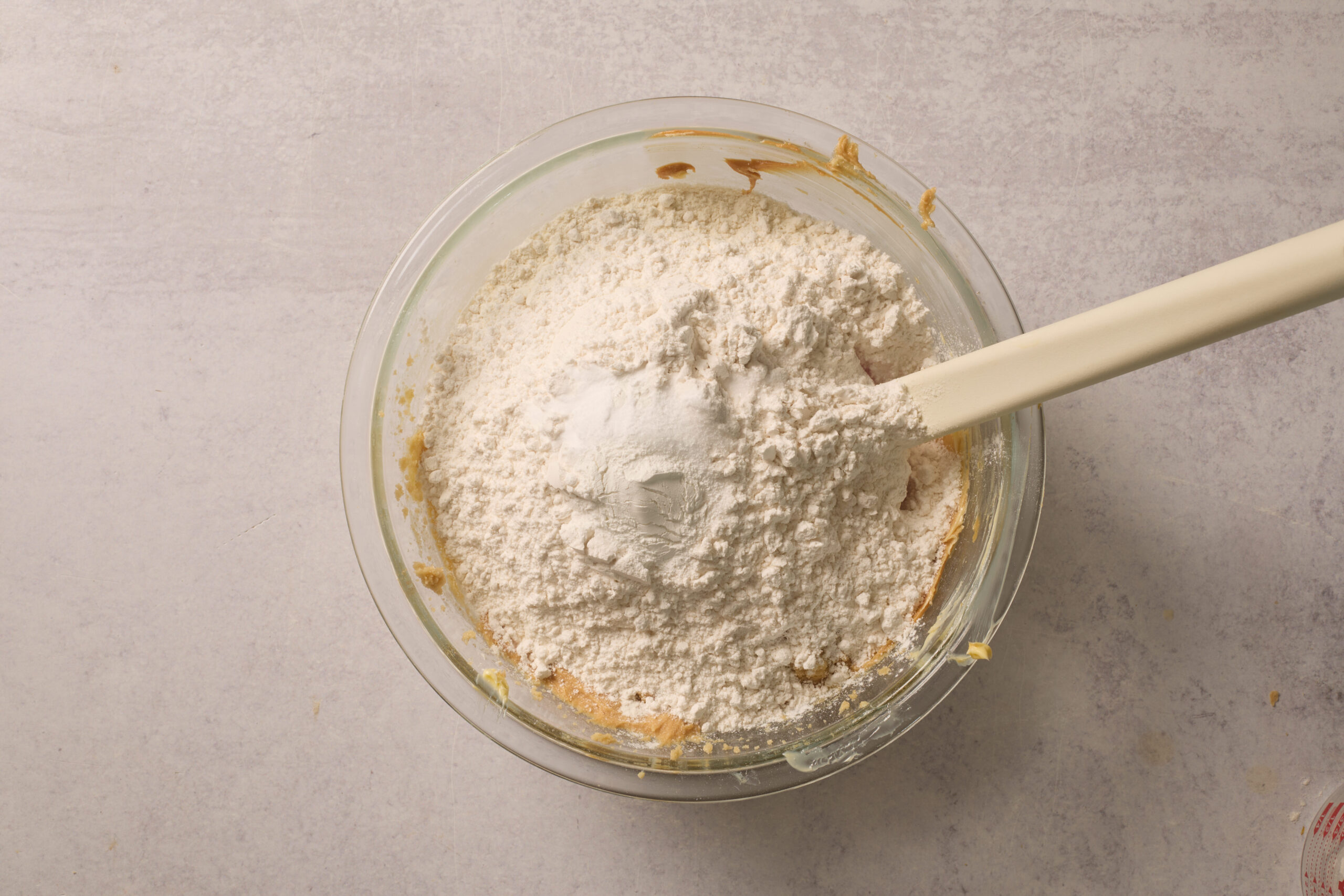 A glass bowl holding flour sits on some half-mixed batter, with a white spatula chilling in the mixture.