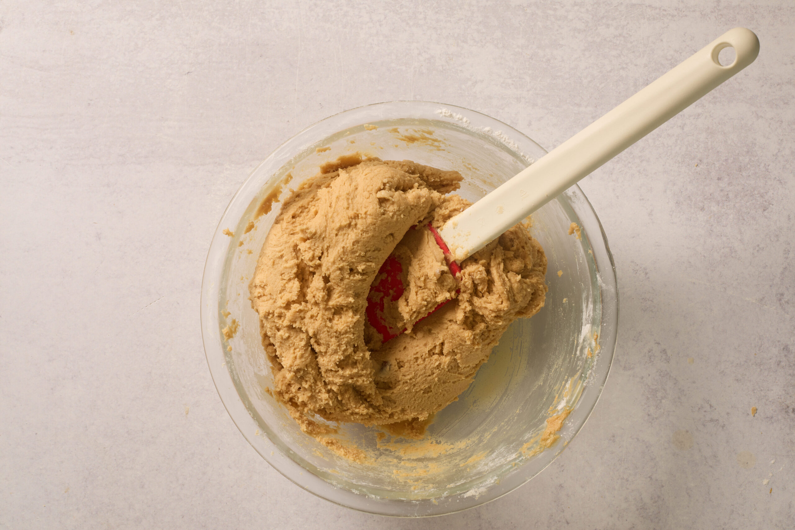Cookie dough getting mixed up in a glass bowl with a white spatula, all sitting on a light countertop.