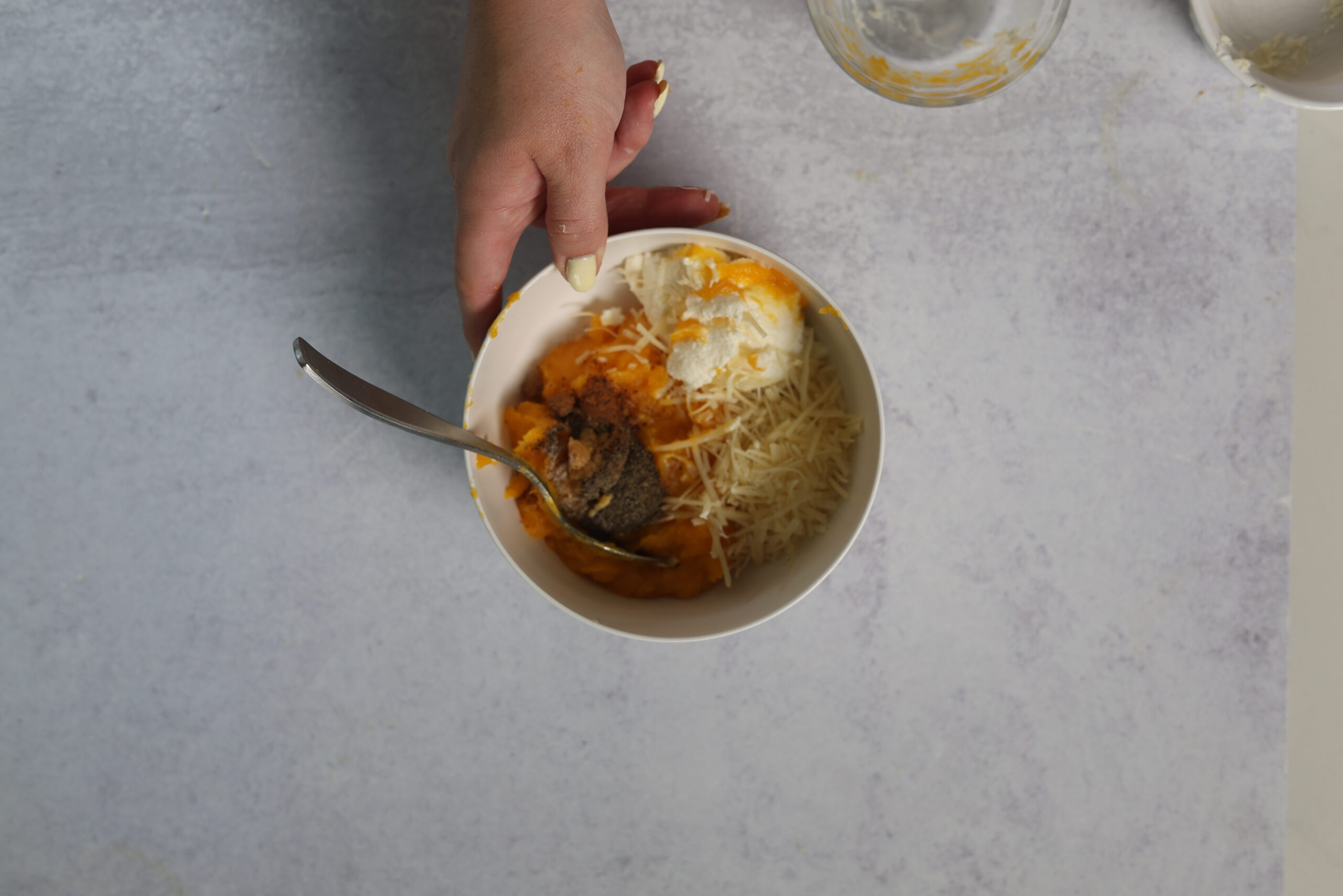 Someone&rsquo;s holding a bowl with ricotta, shredded cheese, pumpkin, and pepper&mdash;ravioli filling ready! Spoon inside, gray backdrop.