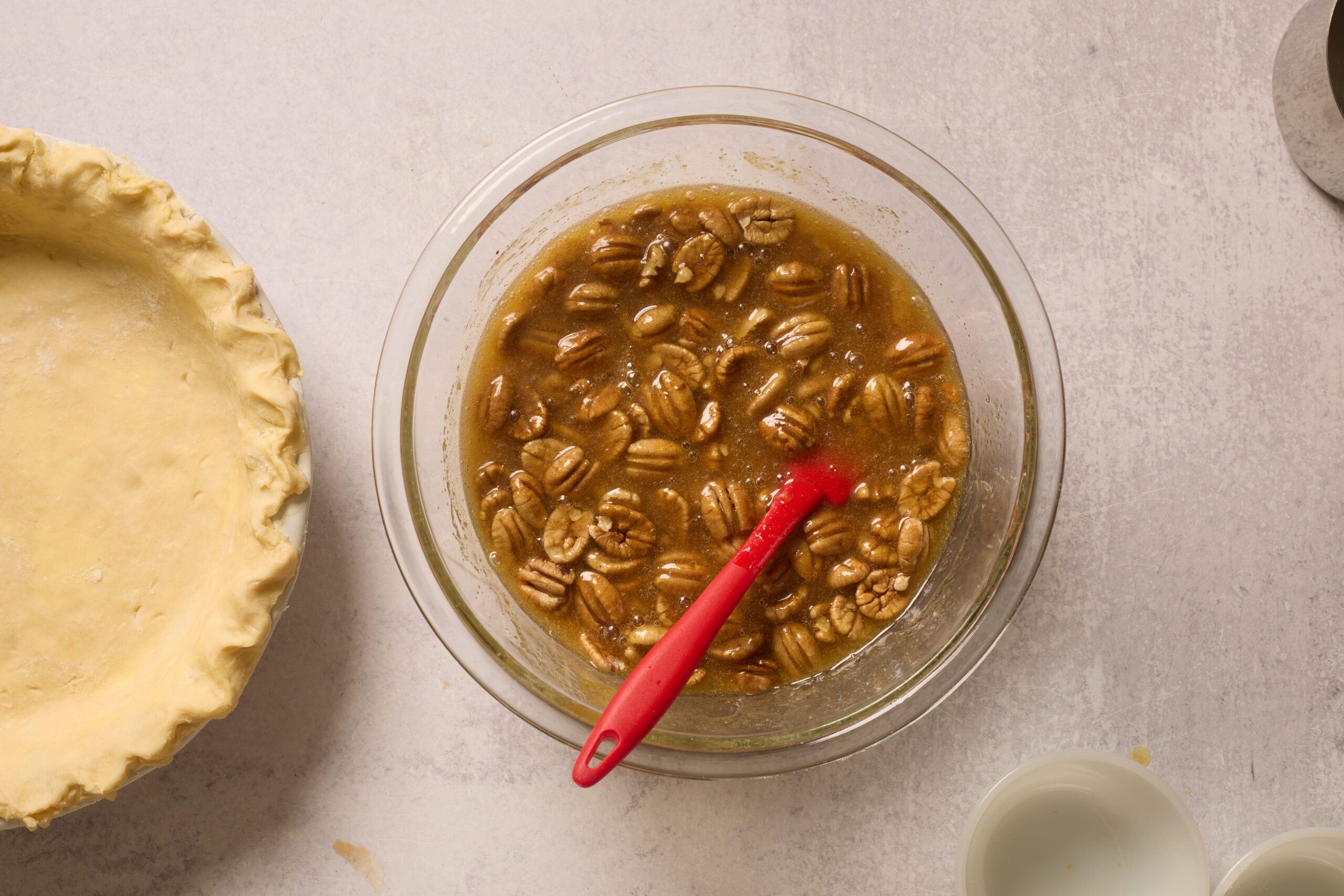 A glass bowl of pecan pie filling with a red spatula is chilling by an unbaked crust on a light countertop.