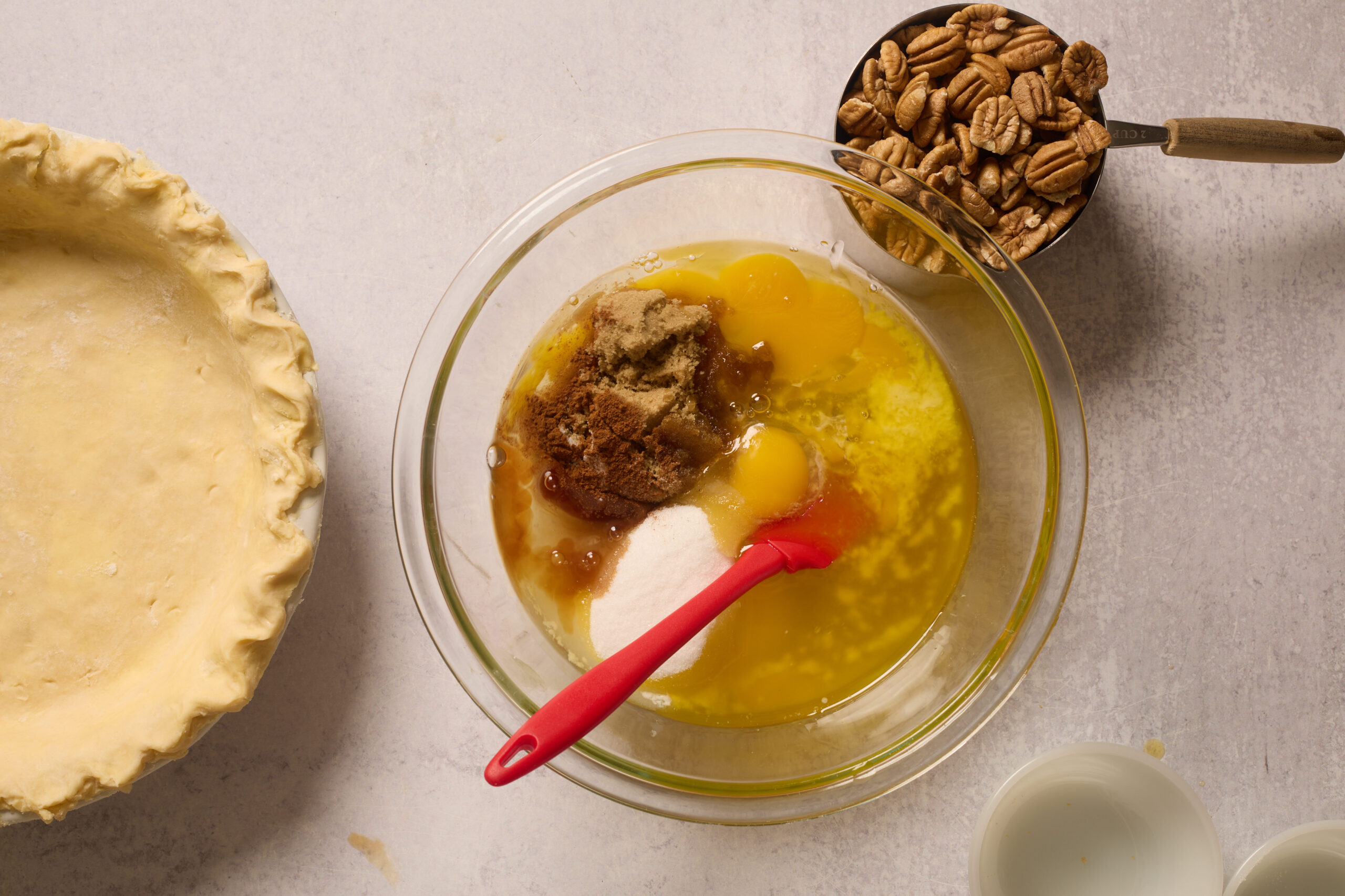 Baking setup: eggs, sugar, and melted butter in a bowl, red spatula, pecans ready to measure, pie crust, and a small bowl of liquid.