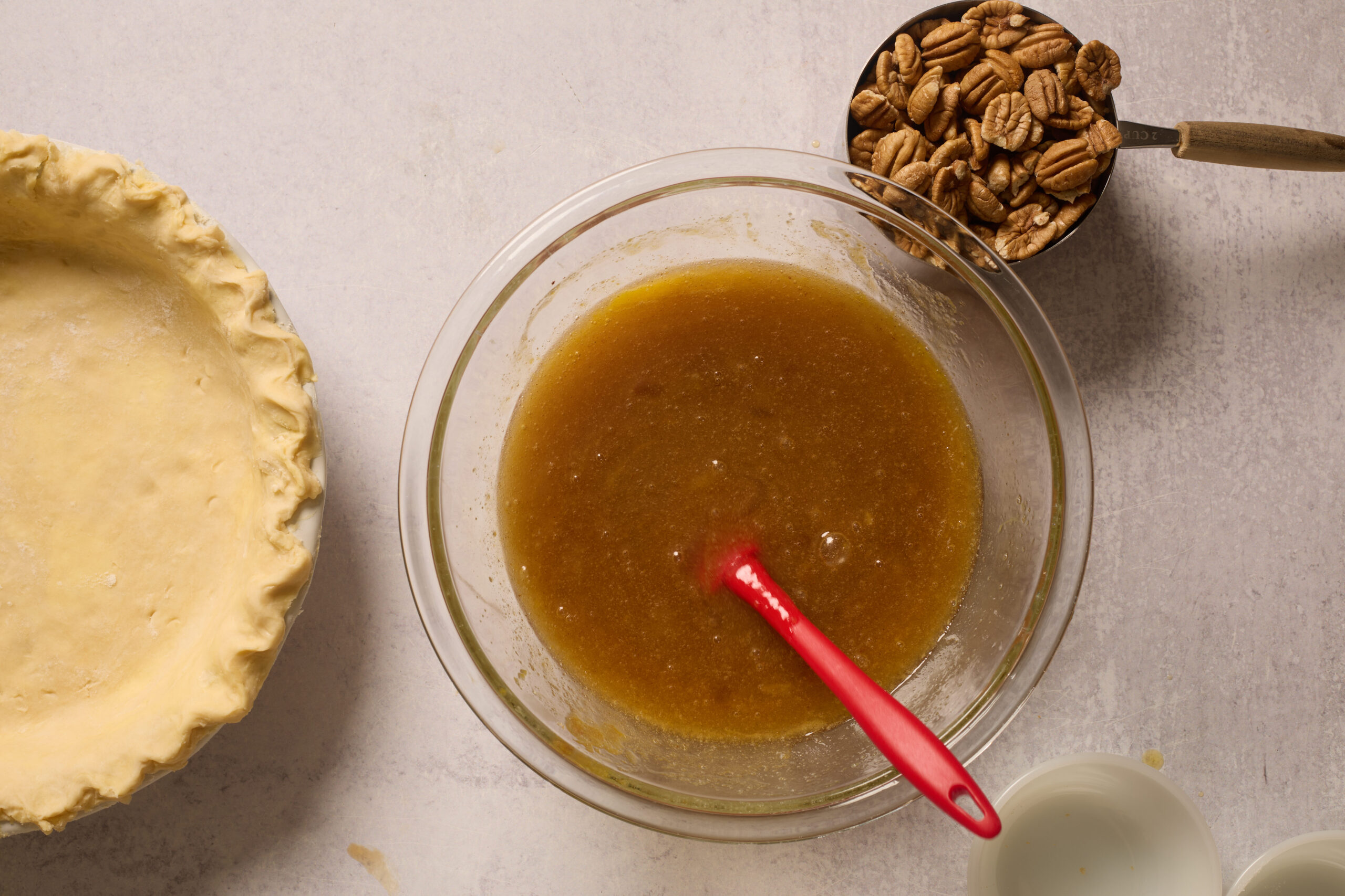 Glass bowl with brown pie filling and a red spatula, pecans in a measuring cup, empty pie crust, and small bowl on the counter.