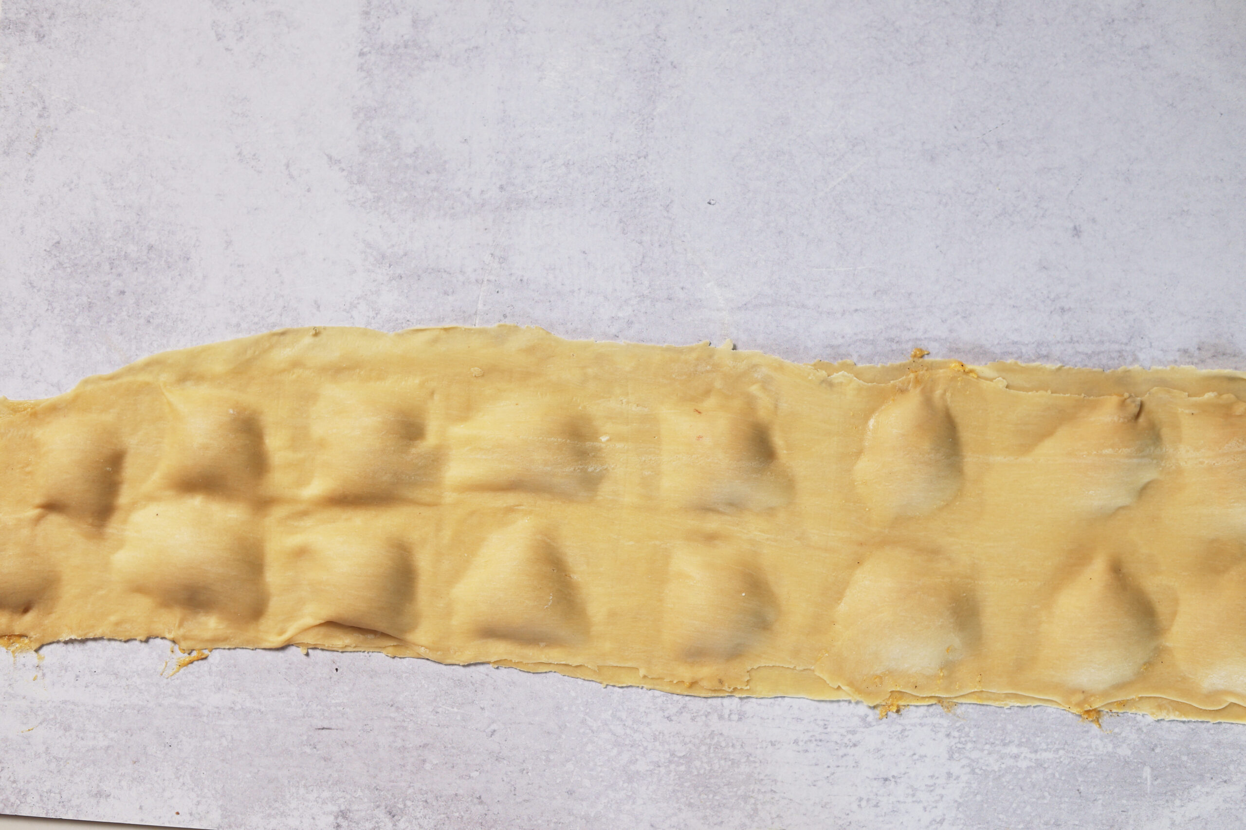 A strip of pasta dough with bumps from ravioli filling sits on a textured surface, ready for a cozy homemade meal.