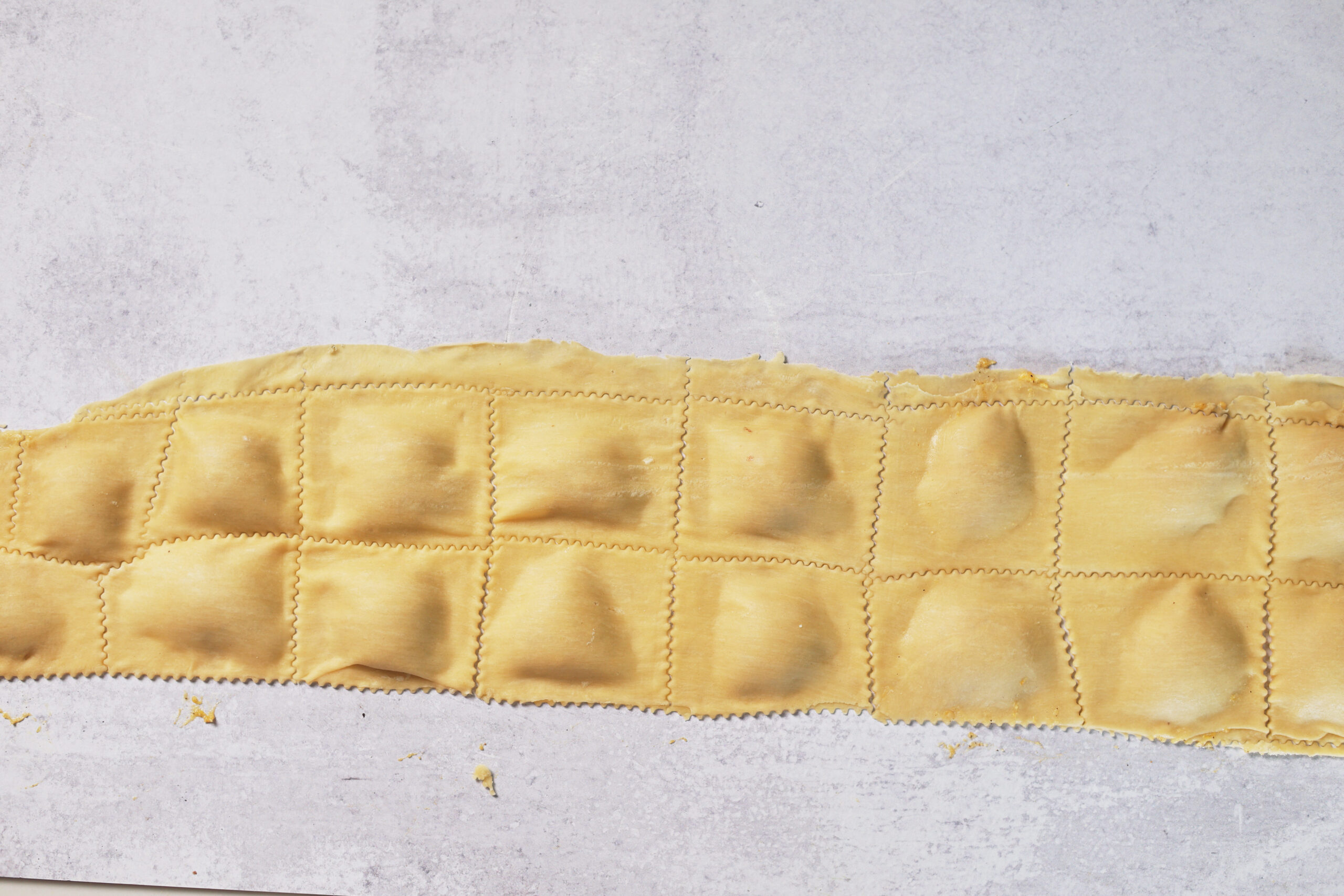 Rolled-out pasta dough with little squares and filling, just waiting to be cut into ravioli on a light-colored surface.