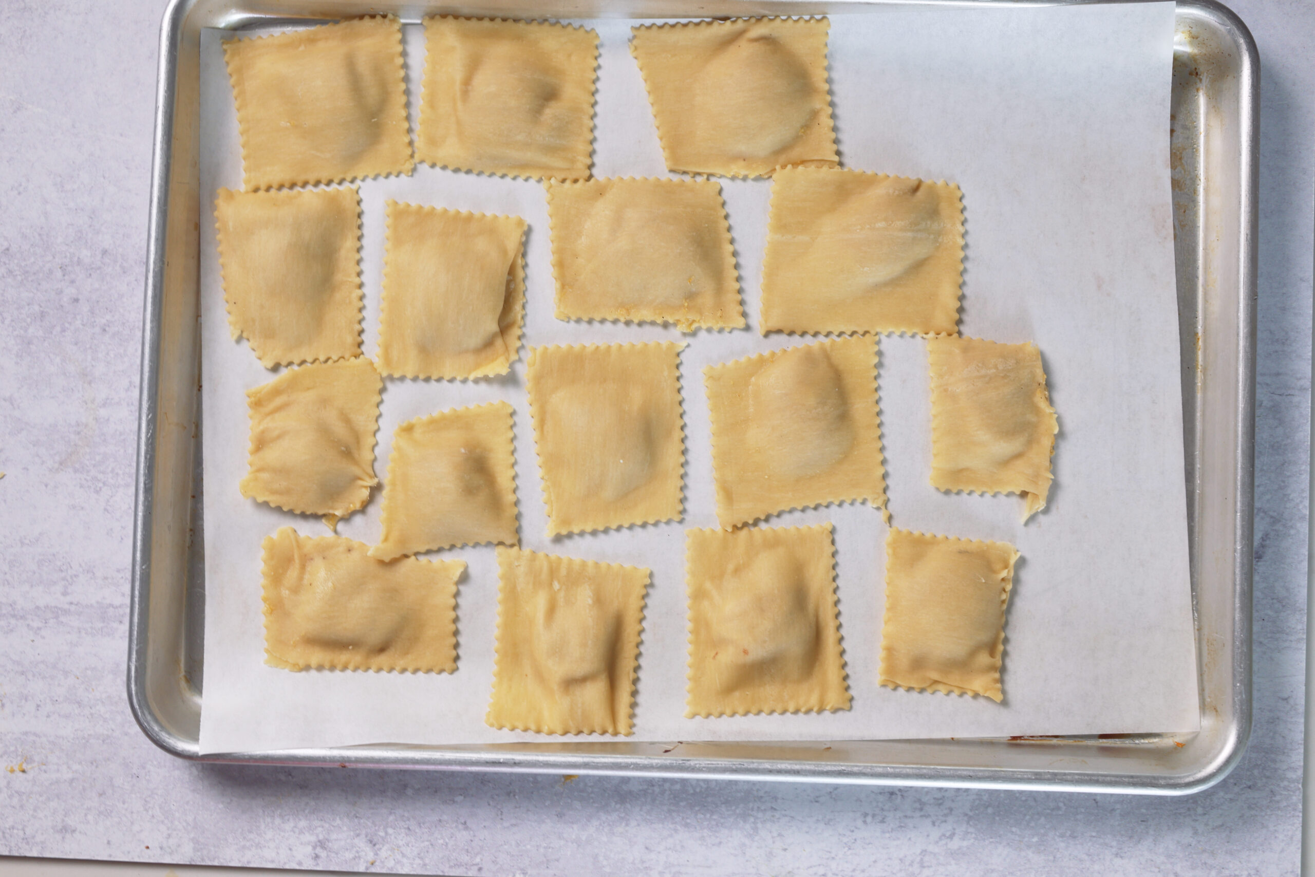 Sixteen homemade ravioli, like butternut squash, rest in rows on a parchment-lined baking sheet, ready to cook.