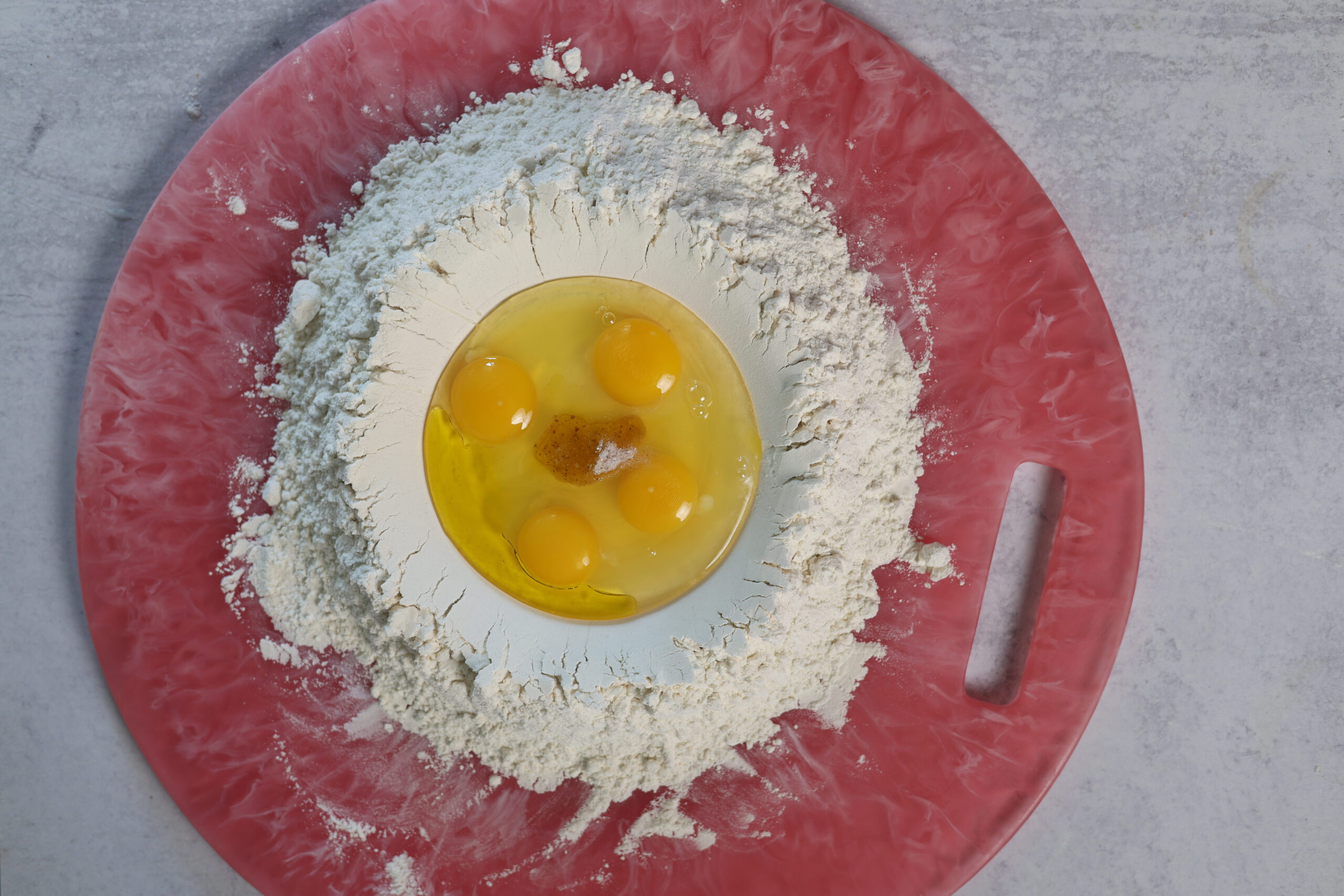 Flour piled up with eggs in the middle and a dash of salt on a red board&mdash;classic pasta prep, ready to get cooking.