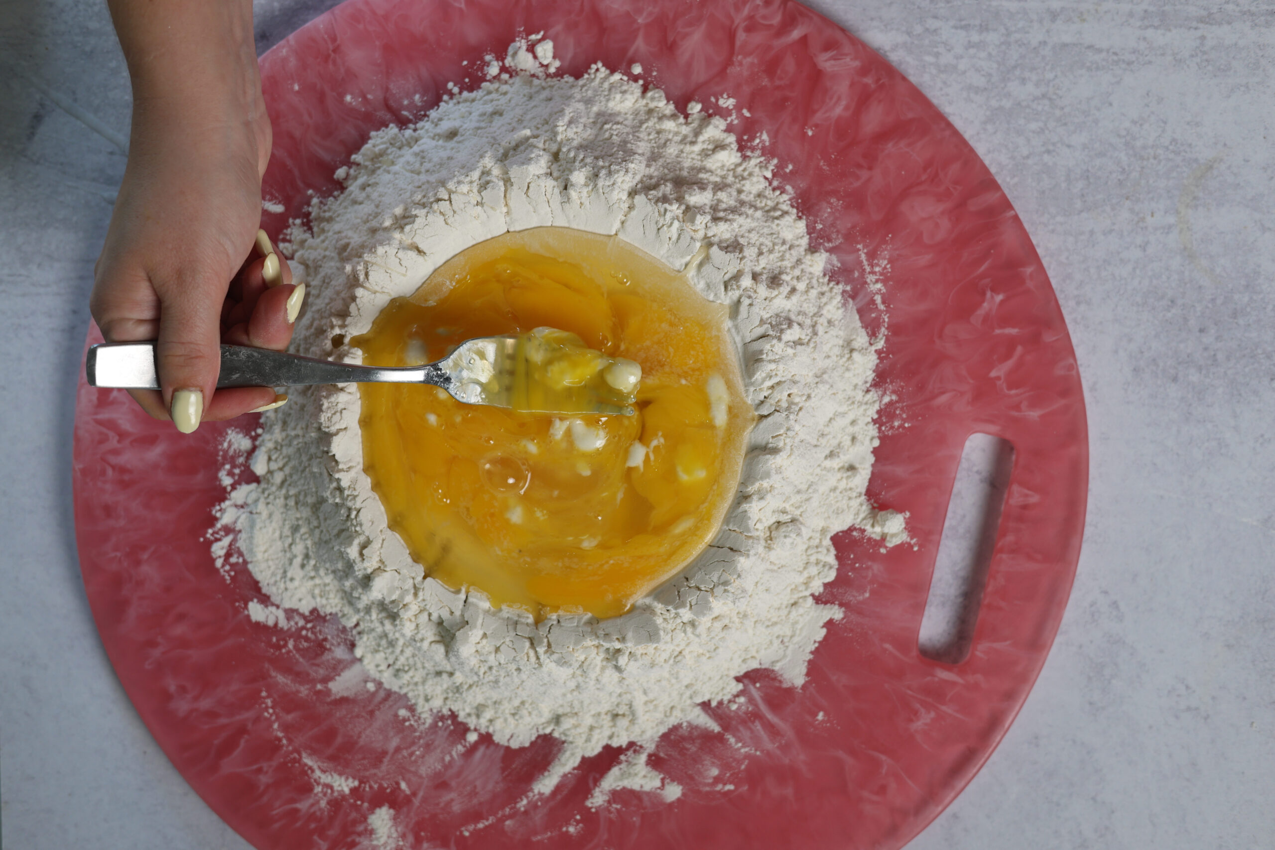 Someone&rsquo;s mixing eggs into a mound of flour with a fork on a pink cutting board, getting ravioli dough started on a gray counter.