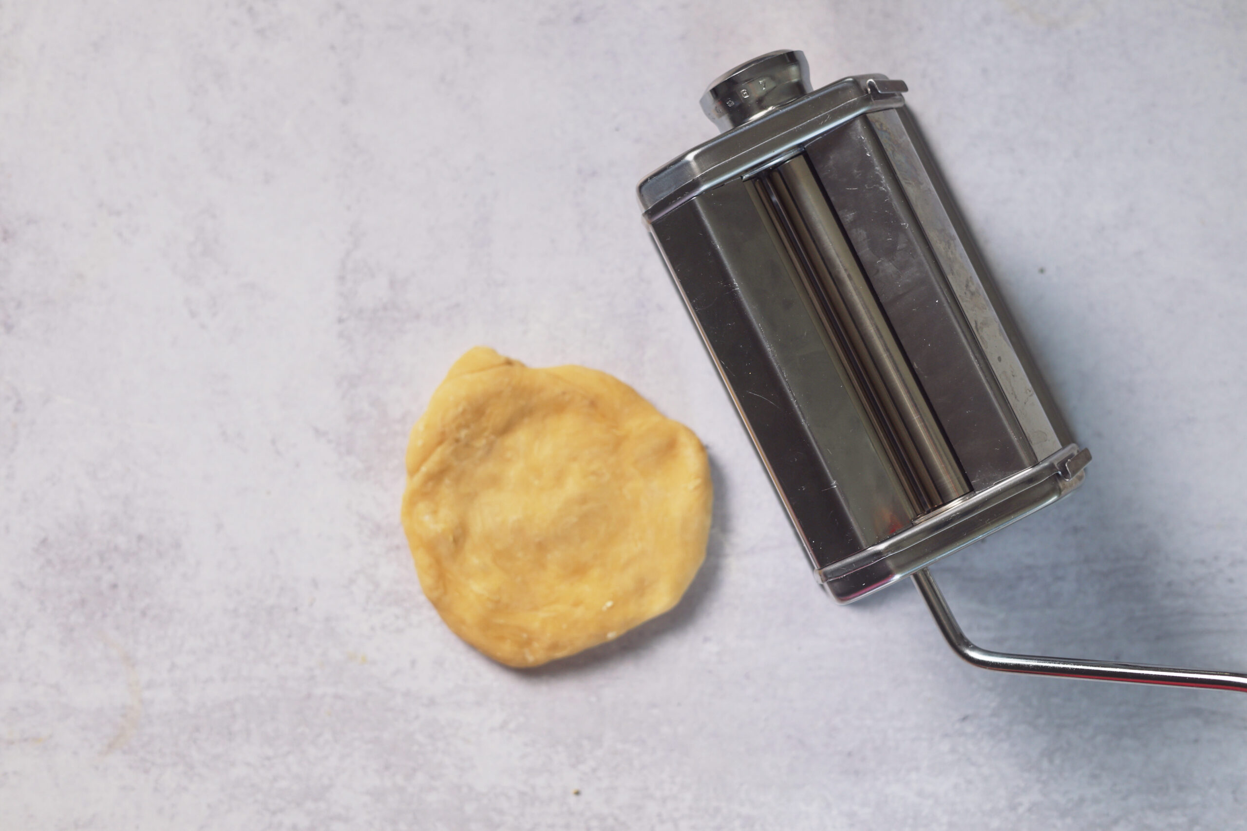 A metal pasta maker sits by a flat dough circle on a light counter, ready for making ravioli or cozy homemade recipes.