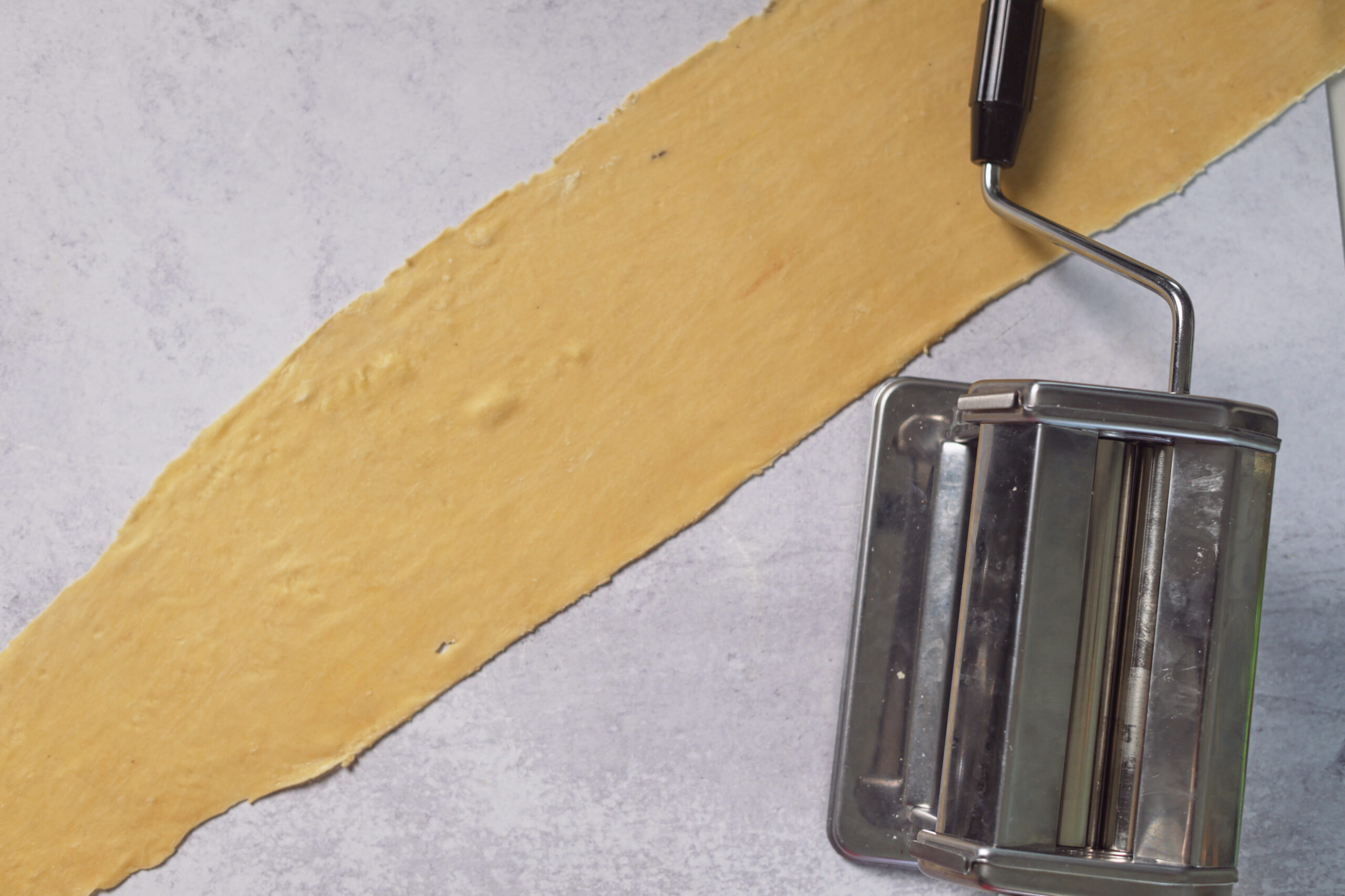 A pasta machine sits beside a long sheet of fresh dough on a light surface, all set for homemade ravioli.