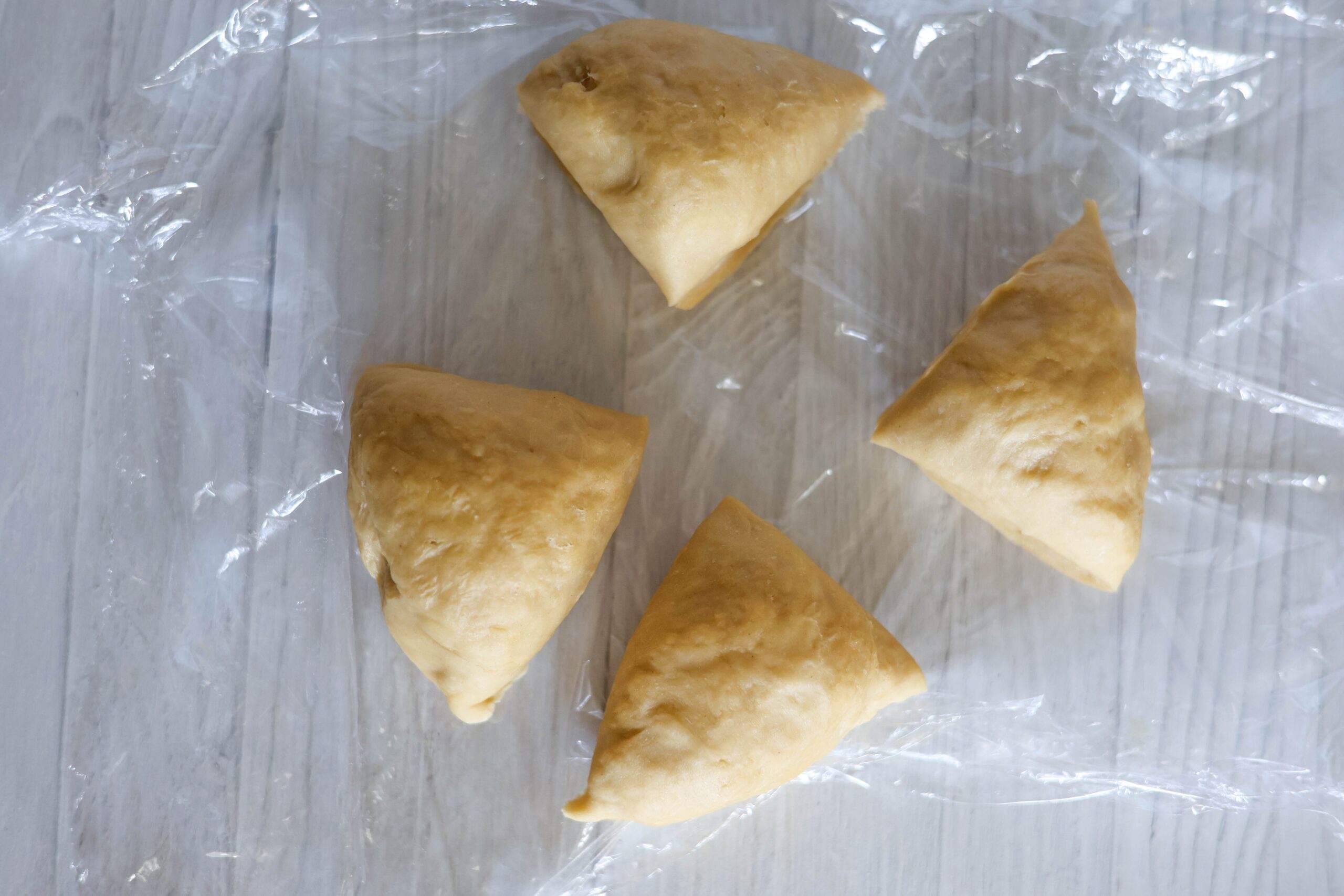 Four triangle-shaped pumpkin ravioli dough pieces sit on plastic wrap over a light wood table.