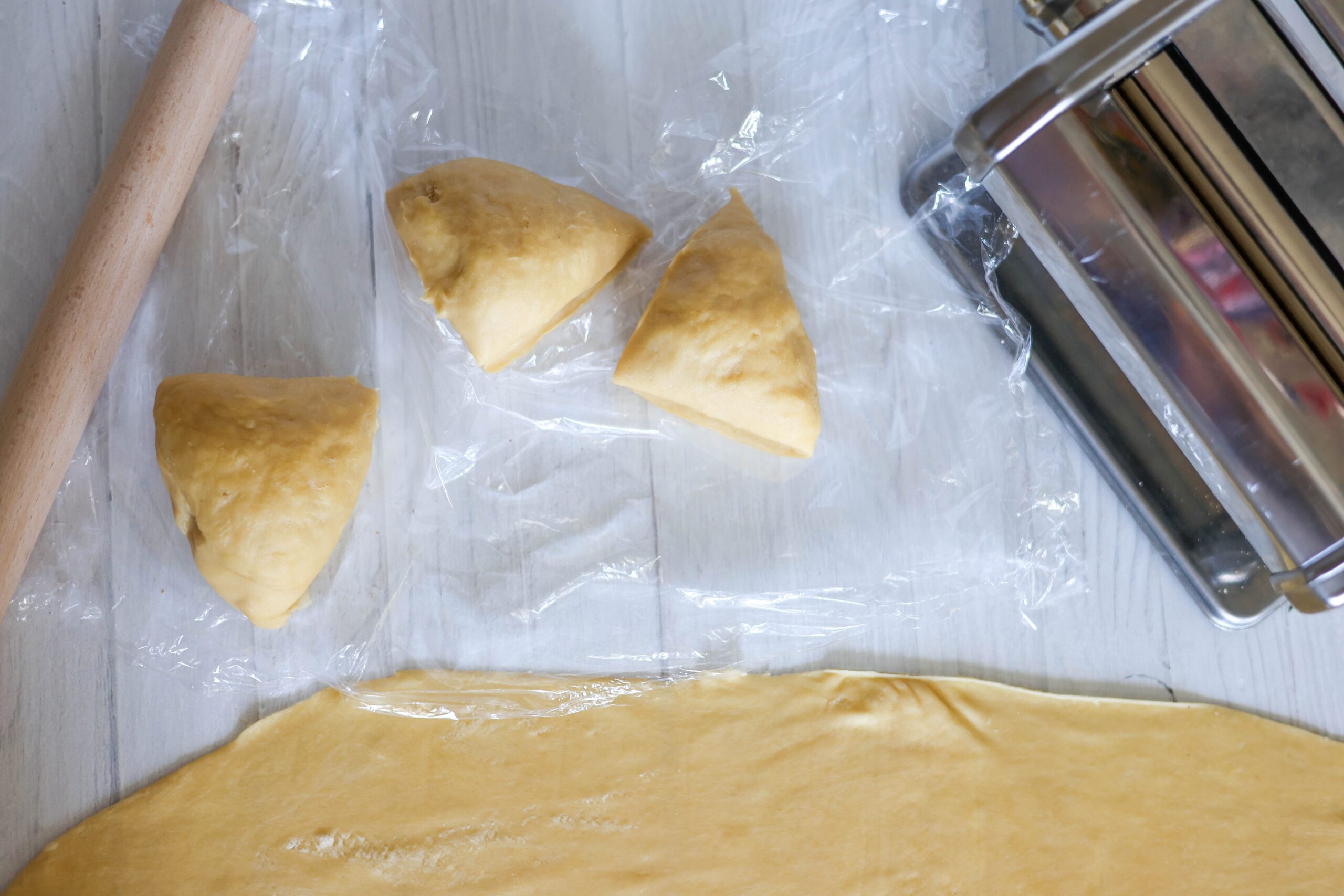 Three dough balls chill on plastic wrap near a rolled-out sheet, ready for pumpkin ravioli. Pasta machine and rolling pin nearby.
