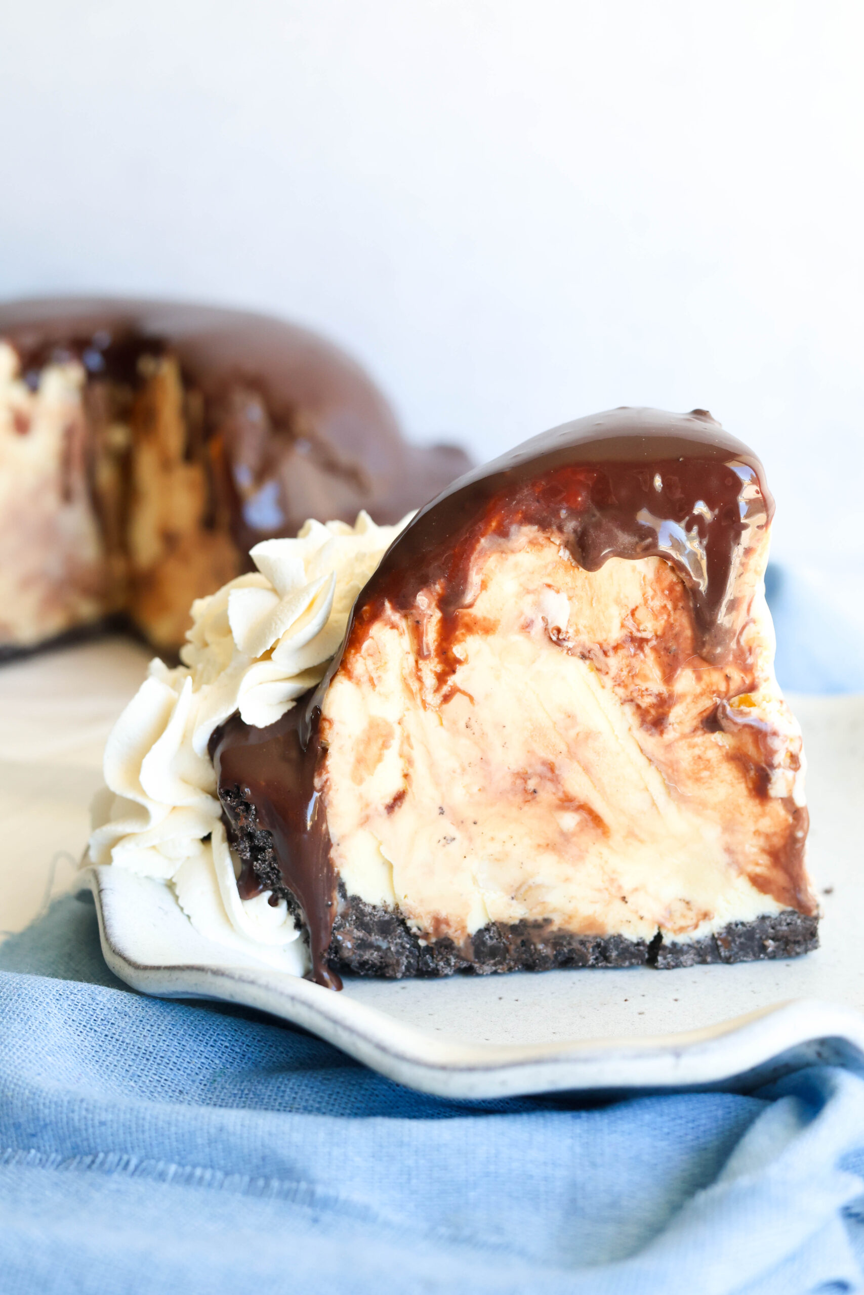 A slice of marble cheesecake with a thick Oreo crust, chocolate ganache, and whipped cream sits on a plate. Cake in back.