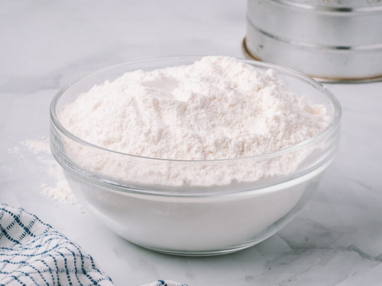 A glass bowl with flour sits on a light marble counter by a blue-and-white checkered cloth, ready for your bread recipe.