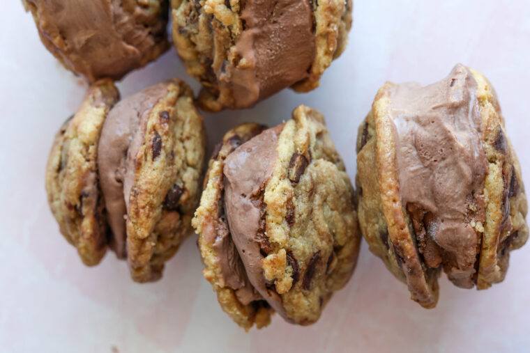 Five chocolate cookie ice cream sandwiches with chocolate ice cream chill on a light pink surface, looking super tasty.