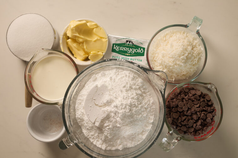 Samoa cookie ingredients&mdash;sugar, butter, cream, coconut flakes, chocolate chips&mdash;lined up on a countertop.