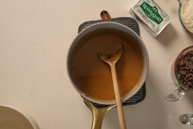 A pan of caramel sauce for Samoa cookies is on a trivet, with a whisk, chocolate chips, coconut, and butter nearby on the counter.