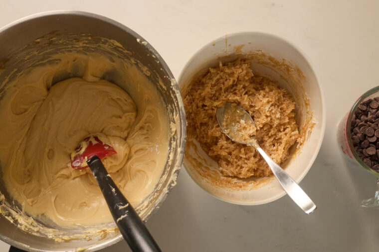Three bowls on the counter&mdash;one has cookie batter with a spatula, another coconut mix with a spoon, and the last has choc chips.