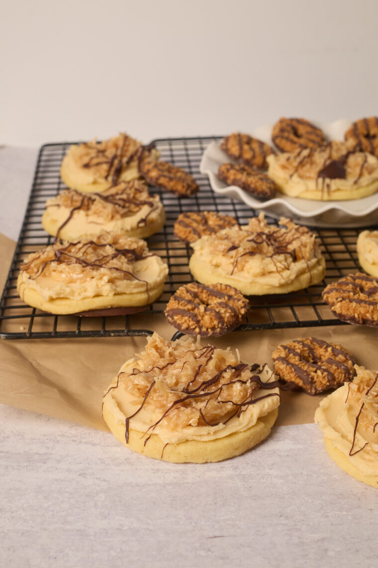 A bunch of frosted samoa cookies topped with caramel, coconut, and chocolate are on a rack, plus some dipped in chocolate nearby.