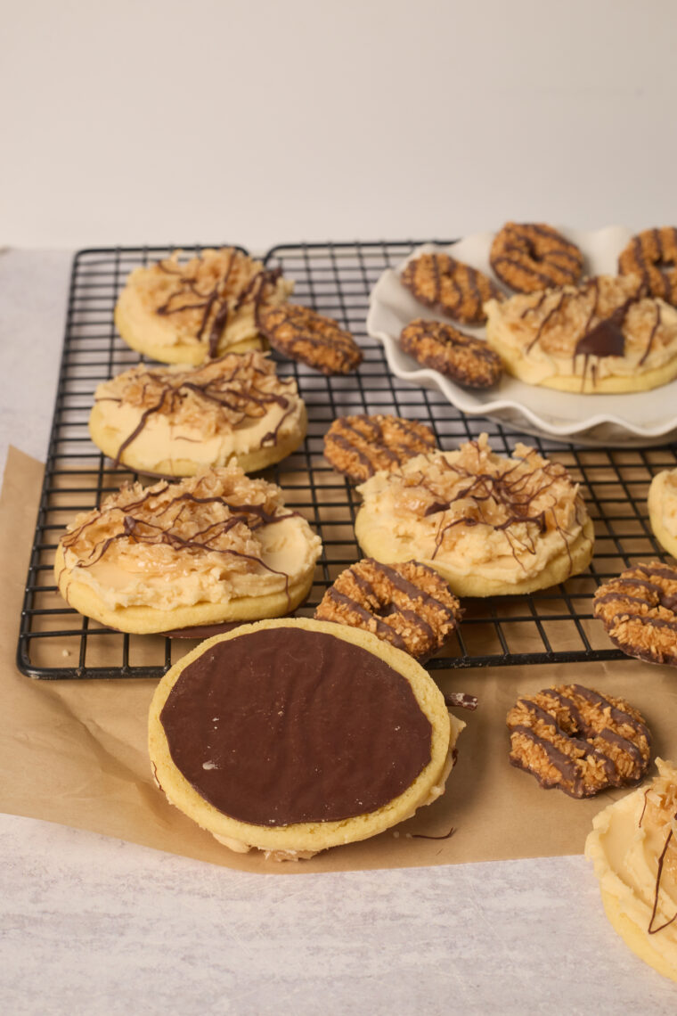 Samoa sugar cookies drizzled with chocolate and caramel chill on a rack, with more cookies and treats nearby on a plate and paper.