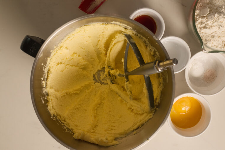 Top-down shot of a bowl with creamed butter and sugar, plus little bowls of vanilla, flour, eggs, and salt nearby on the counter.