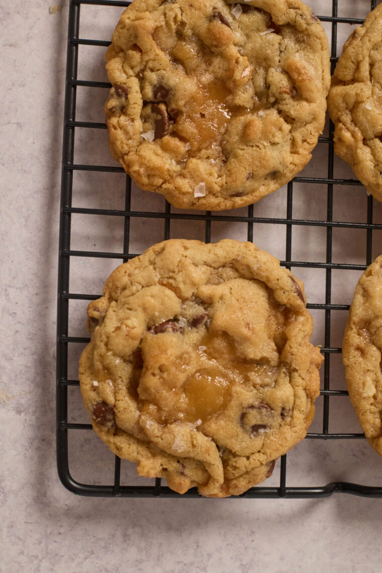 Three chocolate chip cookies with gooey caramel centers cool on a black rack over a light counter, baked to spread just right.